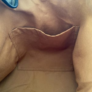 Pu&ograve; includere: Vista interna di una borsa tote in tela. L'interno della borsa &egrave; di un caldo colore marrone, con una tasca abbinata cucita sul davanti. Il bordo superiore della borsa &egrave; di un blu-verde pi&ugrave; scuro, con chiusura a cerniera. La borsa sembra essere realizzata in tela resistente.