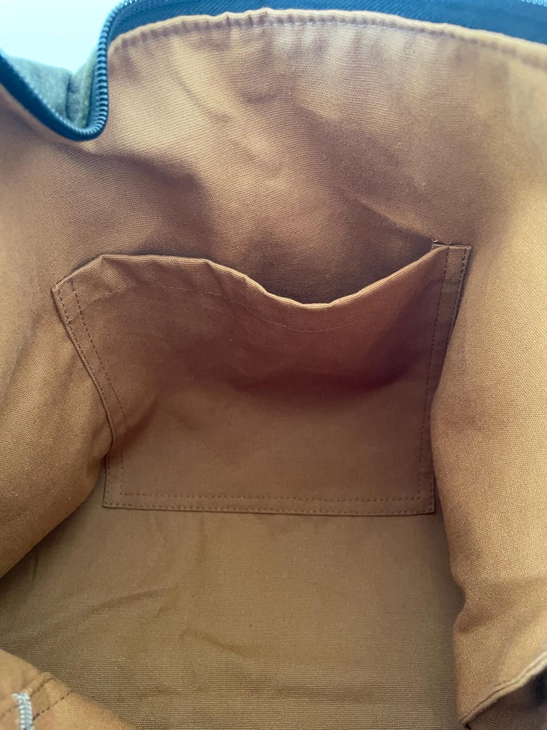 Pu&ograve; includere: Vista interna di una borsa tote in tela. L'interno della borsa &egrave; di un caldo colore marrone, con una tasca abbinata cucita sul davanti. Il bordo superiore della borsa &egrave; di un blu-verde pi&ugrave; scuro, con chiusura a cerniera. La borsa sembra essere realizzata in tela resistente.