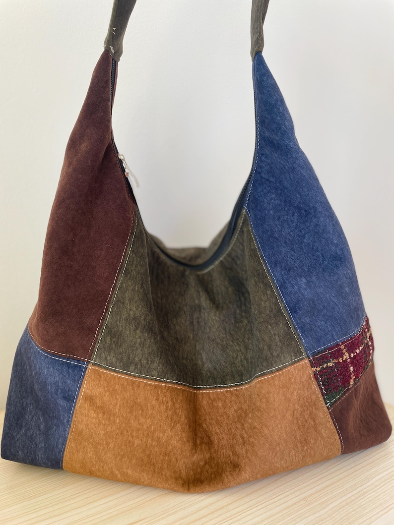 Pu&ograve; includere: Una borsa a tracolla patchwork con una variet&agrave; di colori, tra cui marrone, blu e beige. La borsa ha una consistenza morbida, simile alla pelle scamosciata e una chiusura con cerniera. Il cinturino della borsa &egrave; di colore grigio scuro.