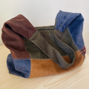 Pu&ograve; includere: Una borsa tote patchwork nei toni del marrone, blu e beige. La borsa presenta una variet&agrave; di trame e colori, con un design cucito. I manici sono marrone scuro e la borsa sembra essere realizzata in pelle scamosciata o materiale simile.