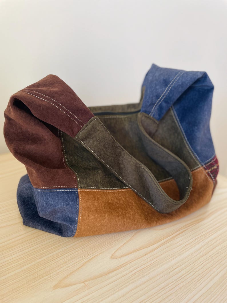 Pu&ograve; includere: Una borsa tote patchwork nei toni del marrone, blu e beige. La borsa presenta una variet&agrave; di trame e colori, con un design cucito. I manici sono marrone scuro e la borsa sembra essere realizzata in pelle scamosciata o materiale simile.