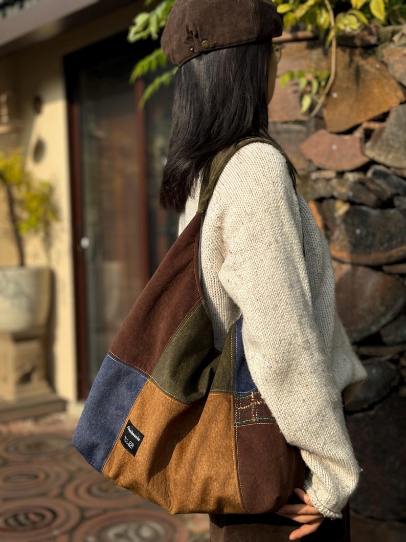 Pu&ograve; includere: Una borsa tote patchwork nei toni del marrone, blu e verde. La borsa presenta una variet&agrave; di trame e motivi, con una piccola etichetta sul davanti. La borsa viene portata a spalla.