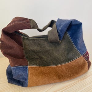 Pu&ograve; includere: Una borsa tote patchwork con una variet&agrave; di colori, tra cui marrone, blu e verde oliva. La borsa ha una consistenza simile alla pelle scamosciata e una chiusura con cerniera. I manici sono di colore marrone scuro.