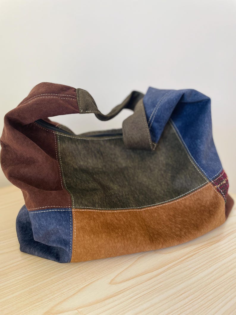 Pu&ograve; includere: Una borsa tote patchwork con una variet&agrave; di colori, tra cui marrone, blu e verde oliva. La borsa ha una consistenza simile alla pelle scamosciata e una chiusura con cerniera. I manici sono di colore marrone scuro.