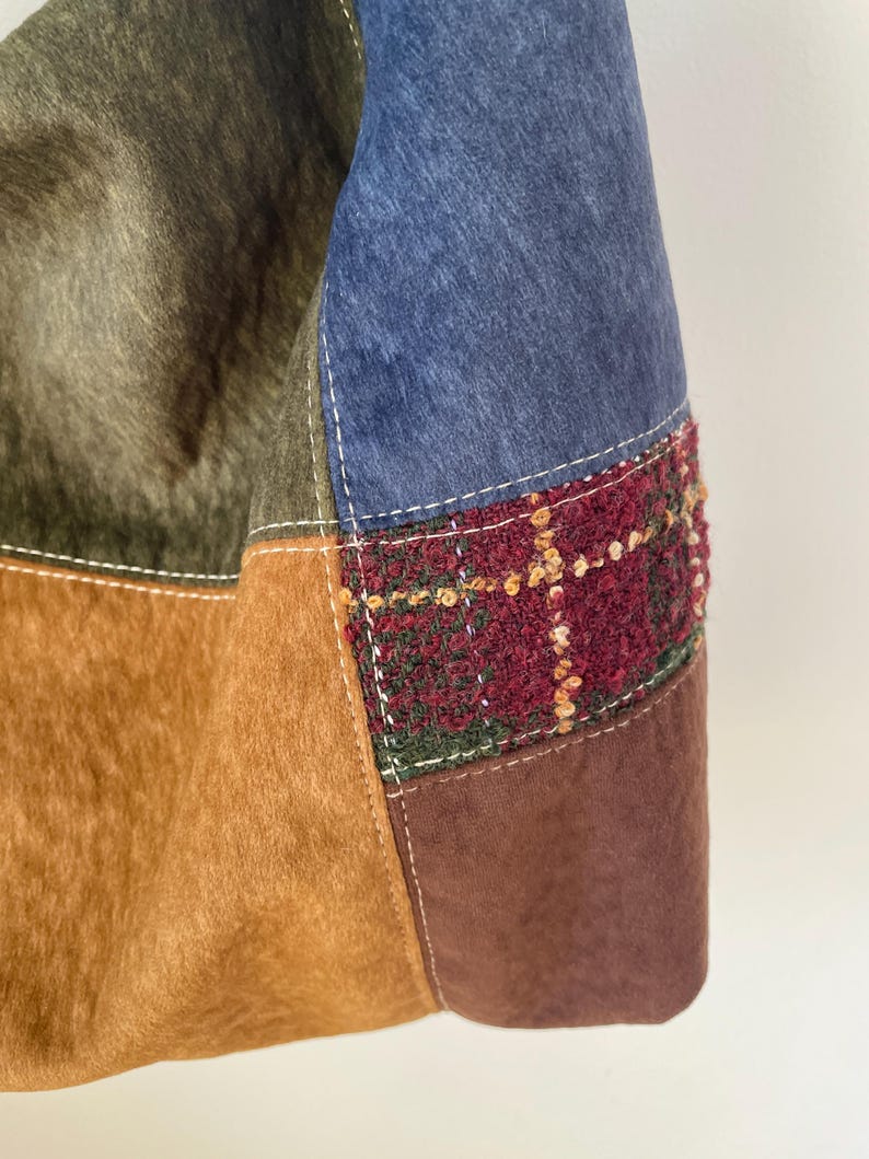 Pu&ograve; includere: Primo piano di una borsa patchwork con varie trame e colori. La borsa incorpora sezioni di tessuti marroni, verde oliva e blu, con un accento tartan strutturato in bordeaux, verde e oro. Le cuciture bianche delineano le cuciture.