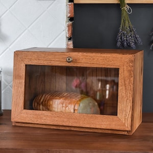 Puede incluir: Una panera de madera con frente de cristal transparente, que muestra pan rebanado. La caja es de madera marrón claro y tiene un pequeño pomo decorativo. Una impresión botánica enmarcada y un tarro con una etiqueta son visibles en el fondo.