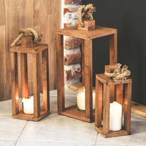 May include: Three wooden lanterns in different sizes, each with a square frame and vertical slats. The lanterns are made of light brown wood and have a rope handle. Lit candles are visible inside, creating a warm glow.
