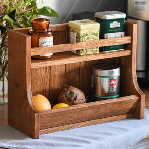 Puede incluir: Un especiero de madera de dos niveles, que contiene varios artículos. El estante superior tiene un frasco de vidrio y dos latas de metal. El estante inferior contiene un coco, una naranja, un limón y una lata de metal. El estante es de madera marrón claro.