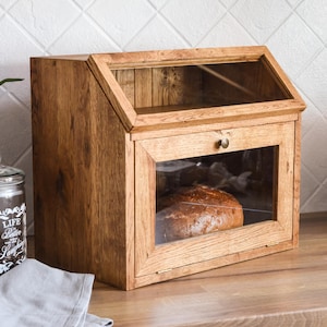 Könnte beinhalten: Holz-Brotdose mit Glasfront und -oberseite. Die Dose ist aus hellbraunem Holz gefertigt und hat einen Klappdeckel. Ein Brotlaib ist im Inneren sichtbar. Die Dose steht auf einer Holz-Arbeitsplatte.