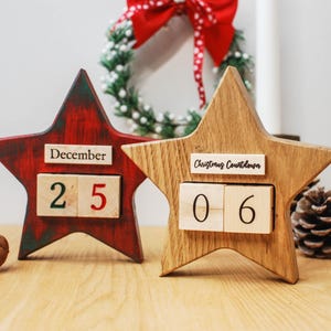 May include: Two wooden star-shaped Christmas countdown calendars. One is red with "December 25", the other is natural wood with "Christmas Countdown 06". Festive holiday decor.