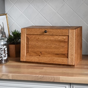 Könnte beinhalten: Eine hölzerne Brotdose mit einer Scharniertür und einem kleinen, runden schwarzen Knopf. Die Box ist aus hellbraunem Holz gefertigt und steht auf einer Holz-Arbeitsplatte. Der Hintergrund zeigt eine weiße geflieste Wand und eine kleine Pflanze.