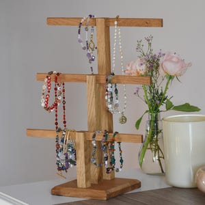 Puede incluir: Un expositor de joyas de madera con múltiples niveles, que contiene varios collares y pulseras. El expositor es de madera clara y presenta un diseño en capas. Se exhiben varios collares y pulseras con cuentas y dijes de colores.