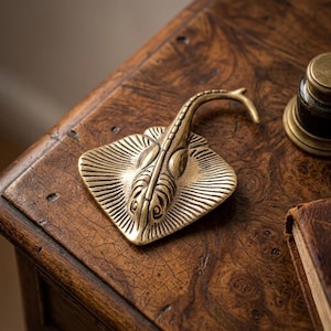 Puede incluir: Un objeto de latón en forma de raya con cola curvada, detallado con líneas grabadas, descansa sobre una superficie de madera oscura. El objeto tiene una estética vintage, con un diseño detallado. Otros objetos de latón son visibles en el fondo.