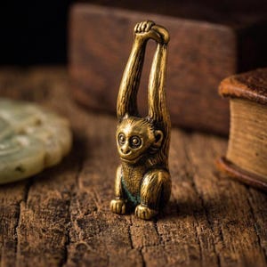 Può includere: Piccola e dettagliata statuetta di scimmia in ottone con le braccia alzate sopra la testa. La scimmia ha un volto sorridente ed è seduta sui suoi quarti posteriori. La statuetta è ambientata su uno sfondo sfocato di legno, che suggerisce un'estetica vintage o antica.
