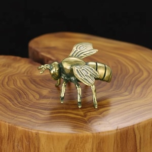 May include: A detailed brass bee figurine with intricate wing and body details. The bee is posed on a wooden surface, showcasing its legs and antennae. The figurine's golden color contrasts with the wood's natural grain.