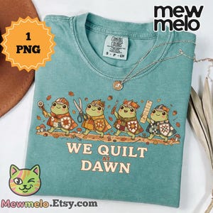 May include: A teal crew neck t-shirt featuring a graphic of four cartoon frogs wearing patchwork capes and holding quilting tools. The text below the frogs reads WE QUILT AT DAWN. The shirt has a Comfort Colors brand label inside the collar.