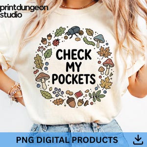 Peut inclure: T-shirt crème avec un motif circulaire. Le motif comprend le texte "CHECK MY POCKETS" entouré d'illustrations de champignons, de feuilles, de glands et d'insectes. Le t-shirt porte également le texte "PNG DIGITAL PRODUCTS".