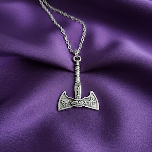 May include: A silver-colored pendant necklace featuring a detailed axe-shaped charm. The charm has intricate designs and is suspended from a delicate chain. The necklace is displayed against a deep purple fabric background.