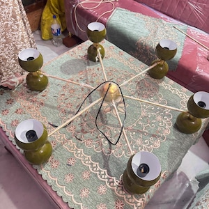May include: A six-arm chandelier featuring olive green spherical shades with white interiors. The fixture has a central gold-toned hub with thin metal arms extending outward, each holding a light socket. A black power cord is attached to the center.