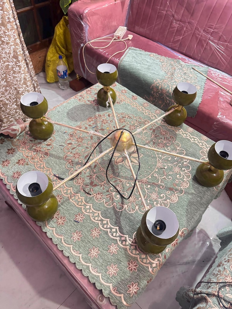 May include: A six-arm chandelier featuring olive green spherical shades with white interiors. The fixture has a central gold-toned hub with thin metal arms extending outward, each holding a light socket. A black power cord is attached to the center.