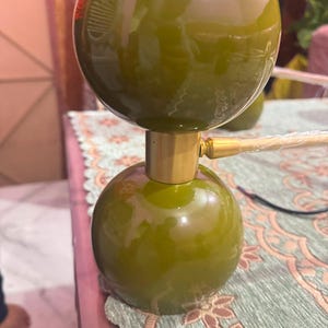 May include: A table lamp featuring two olive green spherical components stacked vertically, separated by a cylindrical gold-toned metal connector. The lamp has a smooth, glossy finish and includes a gold-toned metal arm extending to the right side.