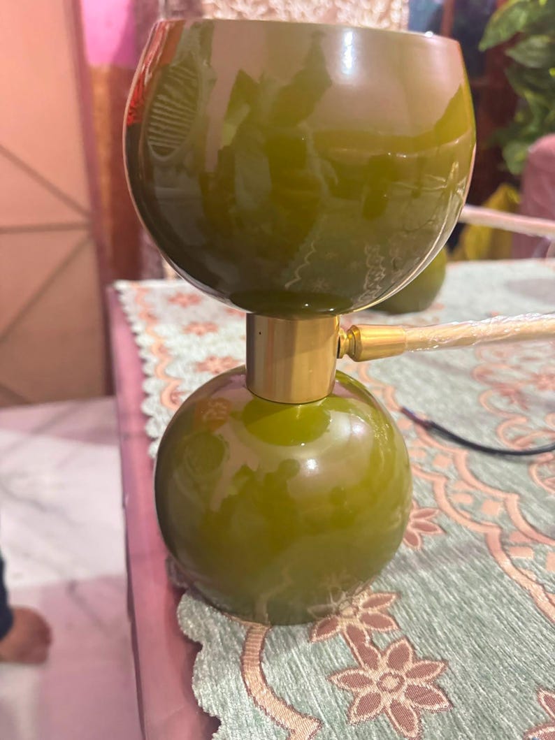 May include: A table lamp featuring two olive green spherical components stacked vertically, separated by a cylindrical gold-toned metal connector. The lamp has a smooth, glossy finish and includes a gold-toned metal arm extending to the right side.