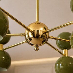 May include: A multi-arm chandelier featuring a central polished gold-tone sphere with radiating arms that extend to support multiple glossy olive green spherical light shades, creating a symmetrical and geometric lighting fixture design.