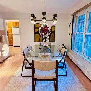 May include: A rectangular glass dining table with a metal frame and six chairs featuring dark wood frames and light-colored upholstered seats. Above the table hangs a multi-light chandelier with dark green globes and a central abstract painting.