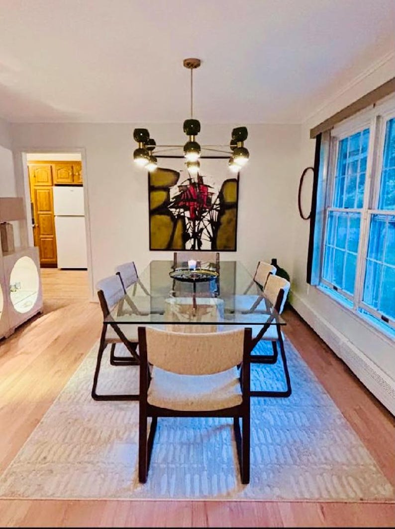 May include: A rectangular glass dining table with a metal frame and six chairs featuring dark wood frames and light-colored upholstered seats. Above the table hangs a multi-light chandelier with dark green globes and a central abstract painting.