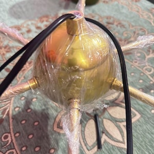 May include: A gold-toned metal light fixture with a central spherical hub and multiple radiating arms. The fixture is partially wrapped in clear protective plastic film and features a black electrical cord extending from the top center.