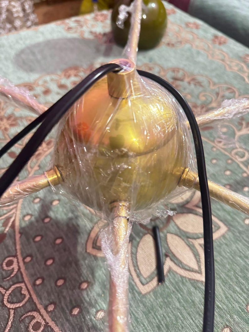 May include: A gold-toned metal light fixture with a central spherical hub and multiple radiating arms. The fixture is partially wrapped in clear protective plastic film and features a black electrical cord extending from the top center.