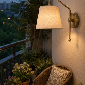 Può includere: Una lampada da parete con braccio in metallo color ottone e paralume conico in tessuto testurizzato bianco sporco. L'apparecchio presenta una piastra circolare a muro e un'asta rivolta verso il basso con un terminale sferico, che emette una luce calda.