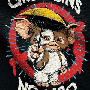 Puede incluir: Diseño gráfico con un Gremlin sosteniendo un paraguas amarillo, con las palabras "GREMLINS" y "NO H2O" en texto blanco. El diseño incluye un círculo rojo y gotas de agua azules sobre un fondo negro.