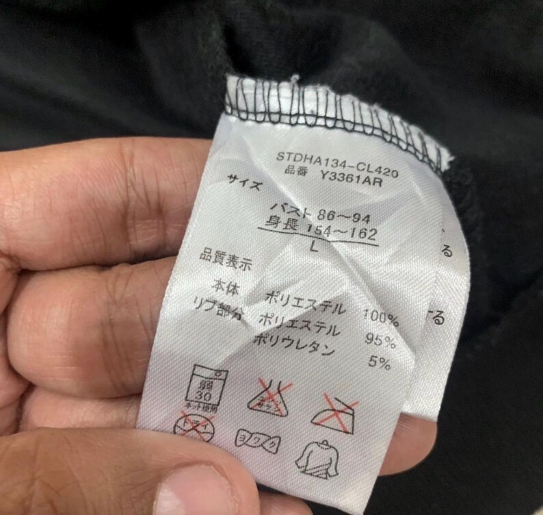Puede incluir: Primer plano de una etiqueta de ropa con texto en japon&eacute;s. La etiqueta incluye detalles sobre la talla, la composici&oacute;n del material (poli&eacute;ster y poliuretano) y las instrucciones de cuidado. La etiqueta es blanca con texto negro.