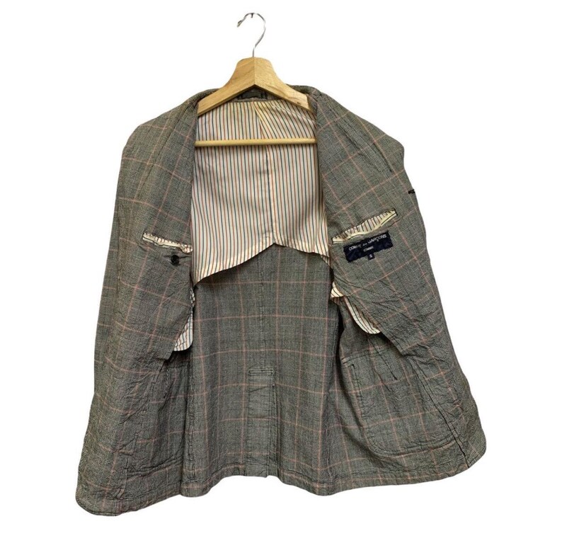 Op de afbeelding: Een grijze blazer met een ruitpatroon, hangend aan een houten hanger. De blazer heeft een gestreepte voering en zichtbare zakken. Het open ontwerp onthult de binnenconstructie en het label.
