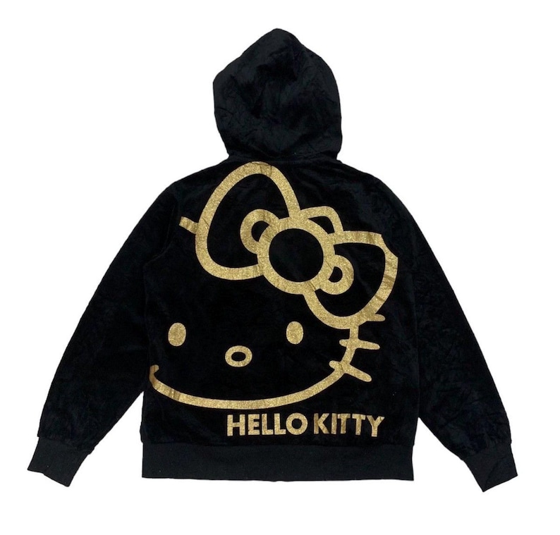 Puede incluir: Sudadera con capucha negra con un dise&ntilde;o de Hello Kitty dorado con purpurina en la espalda. El dise&ntilde;o incluye la cara del personaje y las palabras "Hello Kitty" debajo. La sudadera tiene una cremallera completa y una textura suave y aterciopelada.
