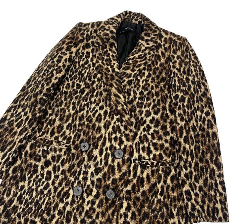 May include: A double-breasted leopard print blazer in shades of brown and black. The blazer features a notched collar, two front pockets, and four dark buttons. The garment is made of a soft, textured fabric.