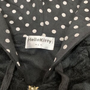 Puede incluir: Sudadera con capucha negra con un estampado de lunares en el interior de la capucha. La etiqueta dice "Hello Kitty" en texto blanco sobre un fondo blanco. La sudadera tiene un cord&oacute;n y una cremallera.