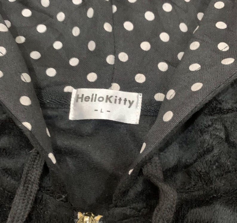 Puede incluir: Sudadera con capucha negra con un estampado de lunares en el interior de la capucha. La etiqueta dice "Hello Kitty" en texto blanco sobre un fondo blanco. La sudadera tiene un cord&oacute;n y una cremallera.