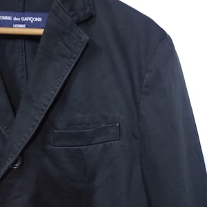May include: A black blazer with a notched lapel and a single chest pocket. The blazer has two buttons and a label that reads "COMME des GAR&Ccedil;ONS HOMME". The jacket is hanging on a wooden hanger.