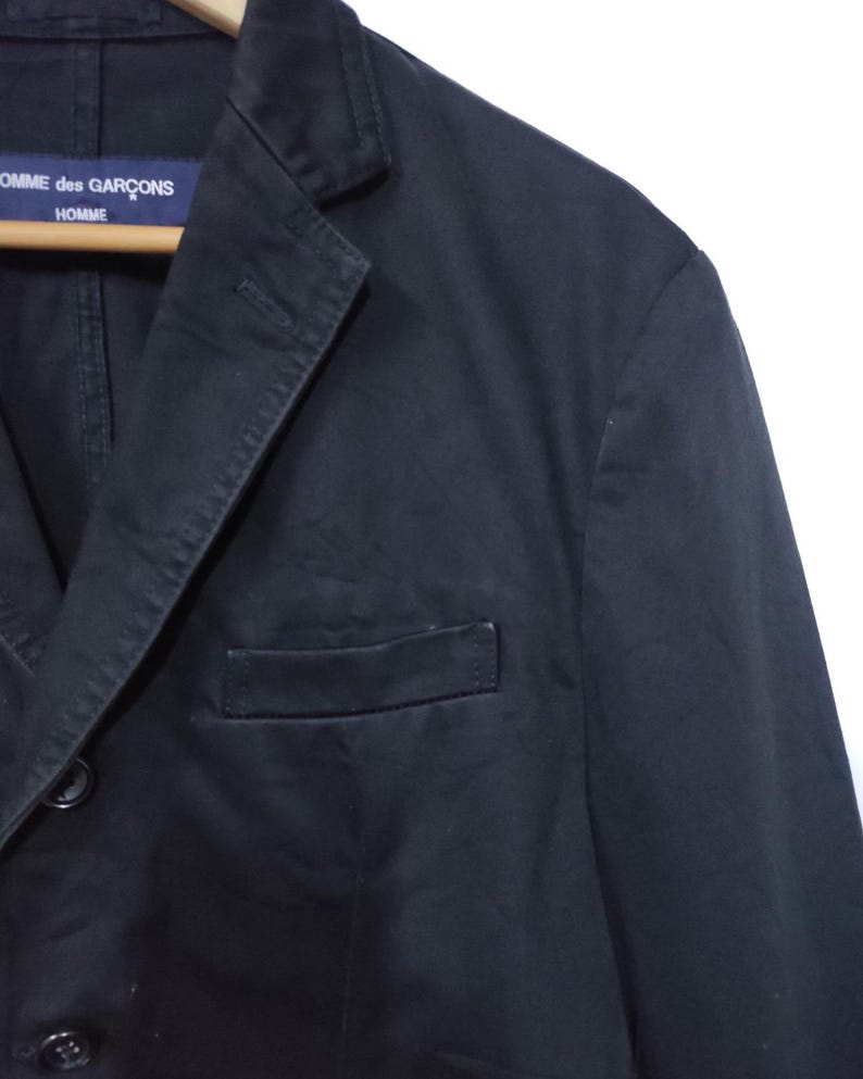 May include: A black blazer with a notched lapel and a single chest pocket. The blazer has two buttons and a label that reads "COMME des GAR&Ccedil;ONS HOMME". The jacket is hanging on a wooden hanger.