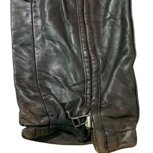 May include: Close-up of a section of a dark brown leather garment. The image shows a zipper with a silver-colored metal pull and a small buckle. The leather has visible stitching and a slightly distressed appearance.
