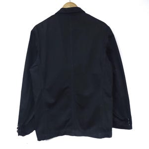May include: A black blazer hanging on a wooden hanger. The blazer has a classic design with a notched lapel, two buttons, and two side pockets. The back of the blazer is visible, showing a tailored fit.