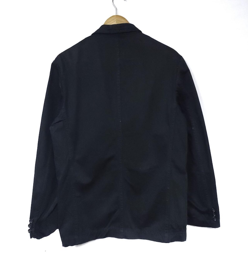 May include: A black blazer hanging on a wooden hanger. The blazer has a classic design with a notched lapel, two buttons, and two side pockets. The back of the blazer is visible, showing a tailored fit.
