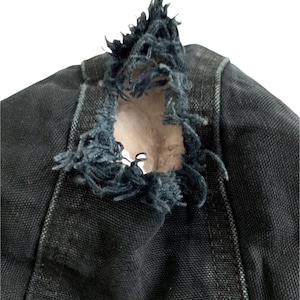 May include: Close-up of a distressed black denim garment. The fabric is torn, revealing a lighter interior. Frayed edges and visible stitching add to the worn aesthetic. The garment appears to be part of a larger piece of clothing.