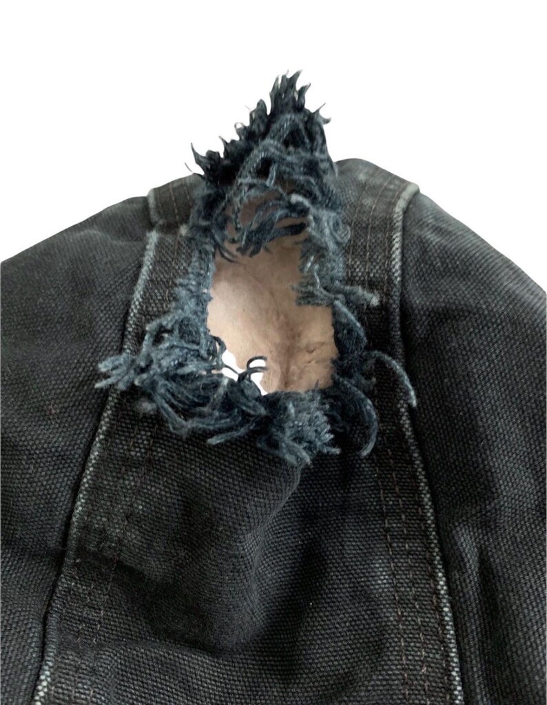 May include: Close-up of a distressed black denim garment. The fabric is torn, revealing a lighter interior. Frayed edges and visible stitching add to the worn aesthetic. The garment appears to be part of a larger piece of clothing.