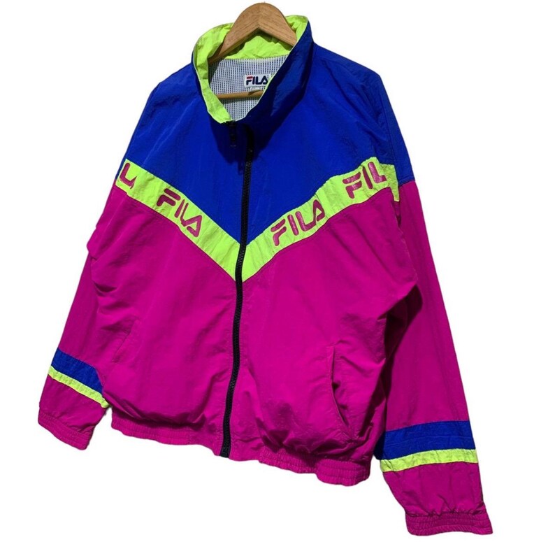 May include: A vintage Fila track jacket in a vibrant color block design. The jacket features a combination of blue, pink, and neon green. The Fila logo is prominently displayed across the chest and sleeves. It has a full zip closure and a stand-up collar.