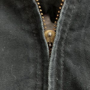 May include: Close-up of a partially zipped, dark gray garment. The zipper is a brass color, with a visible pull tab. The fabric has a textured appearance, and the stitching is visible along the seam.