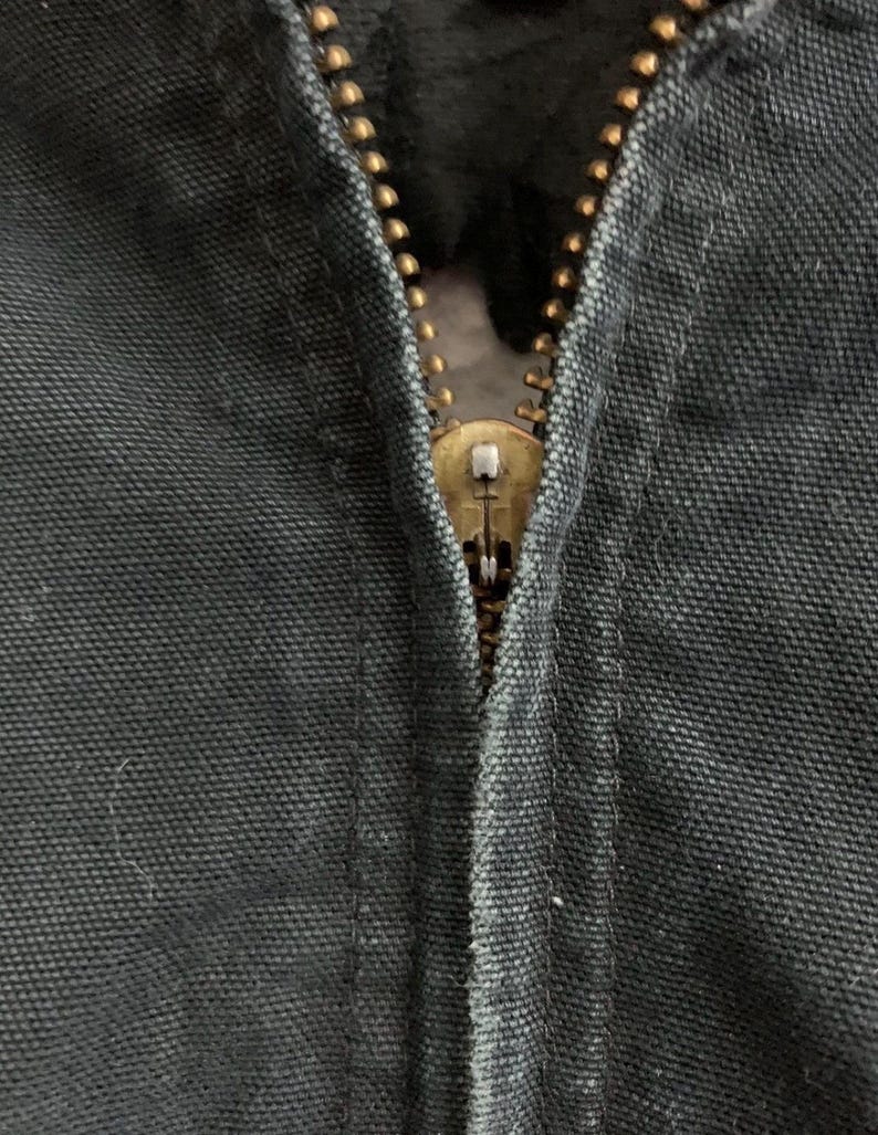 May include: Close-up of a partially zipped, dark gray garment. The zipper is a brass color, with a visible pull tab. The fabric has a textured appearance, and the stitching is visible along the seam.