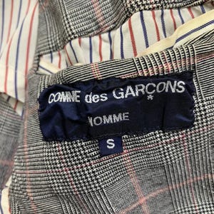 Op de afbeelding: Detail van een blazerinterieur met een zwart, wit en rood ruitpatroon. De voering heeft rode en blauwe strepen op een witte achtergrond. Een marineblauw label leest "COMME des GARCONS HOMME" met een maat "S" tag.